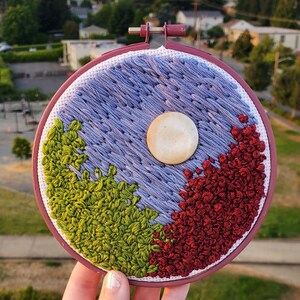 May include: A hand-stitched embroidery hoop with a purple, green, and red design. The design features a white circle in the center and resembles a yin-yang symbol.