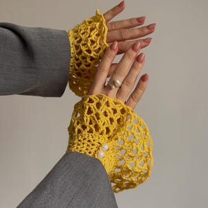 May include: Bright yellow, open-weave crochet wrist cuffs with pearl accents. The cuffs are worn over the sleeves of a gray blazer. A silver ring with a pearl is visible on one hand. The background is a neutral color.