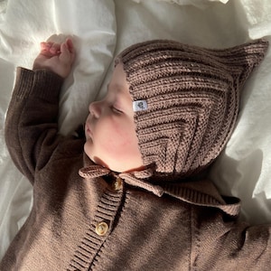 May include: A sleeping baby wearing a brown knitted cardigan and matching bonnet with a pointed top. The cardigan has button closures and ribbed detailing. The baby is lying on a white sheet.