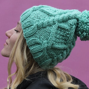 Puede incluir: Un gorro de punto azul turquesa con un gran pompón en la parte superior. El gorro tiene un patrón de punto de cable.