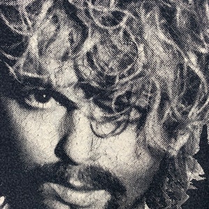 May include: Black and white close-up image of a man's face with long, curly hair. The image is printed on a black t-shirt.