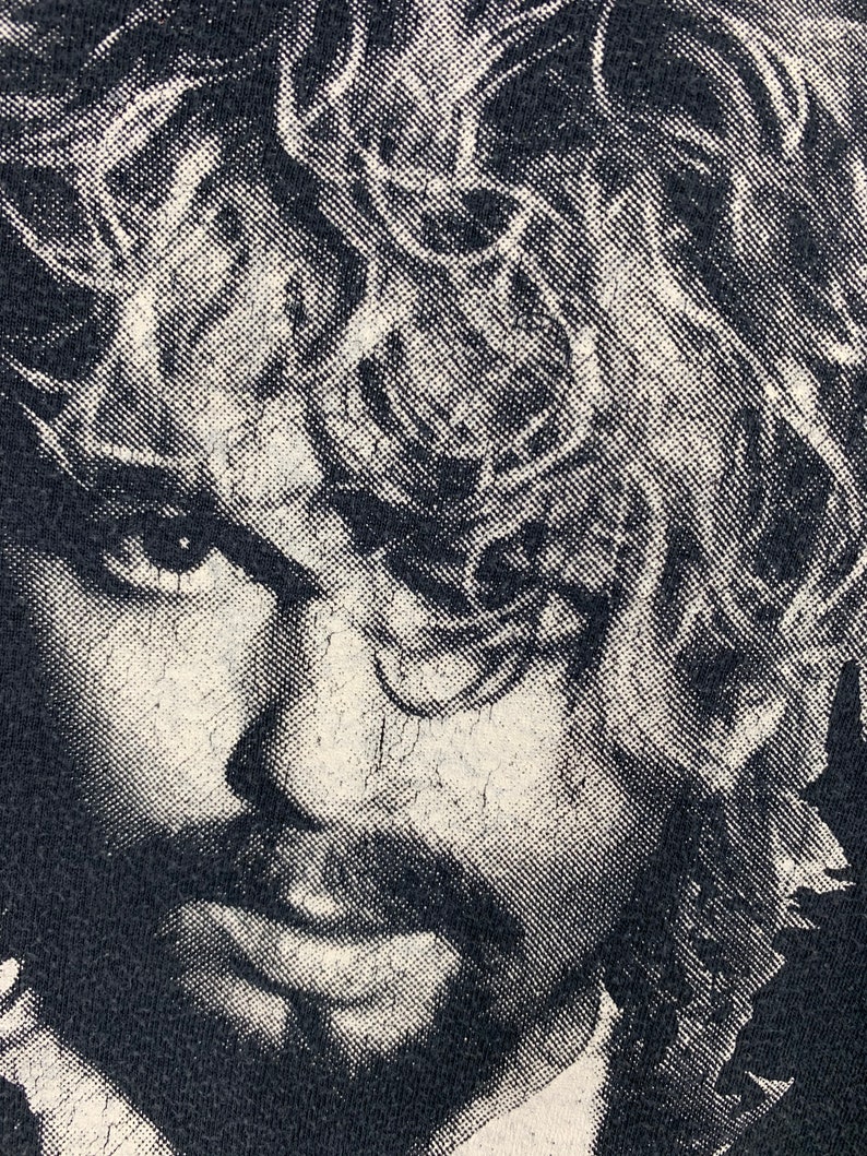 May include: Black and white close-up image of a man's face with long, curly hair. The image is printed on a black t-shirt.