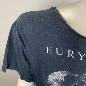 May include: A dark grey t-shirt with a white print of the band Eurythmics. The print features a black and white image of the band's lead singer, Annie Lennox.