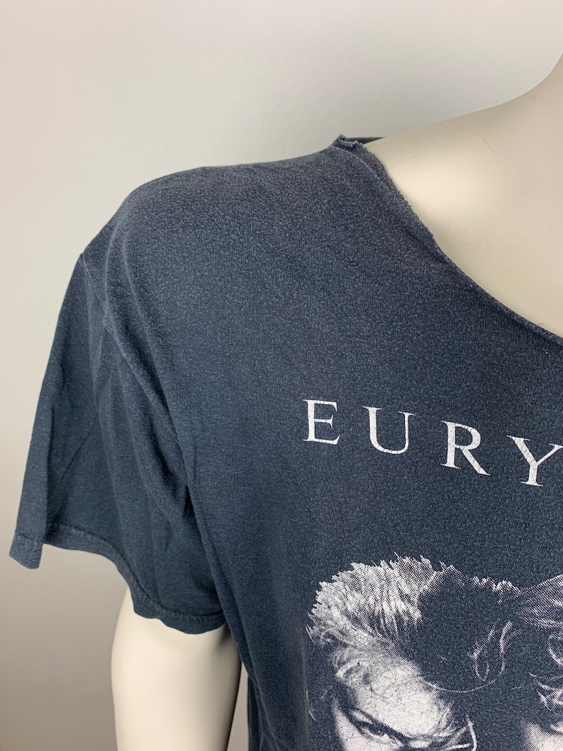 May include: A dark grey t-shirt with a white print of the band Eurythmics. The print features a black and white image of the band's lead singer, Annie Lennox.