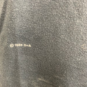 May include: Close-up of a dark grey fabric with the text "&copy; 1986 DnA" printed in white.