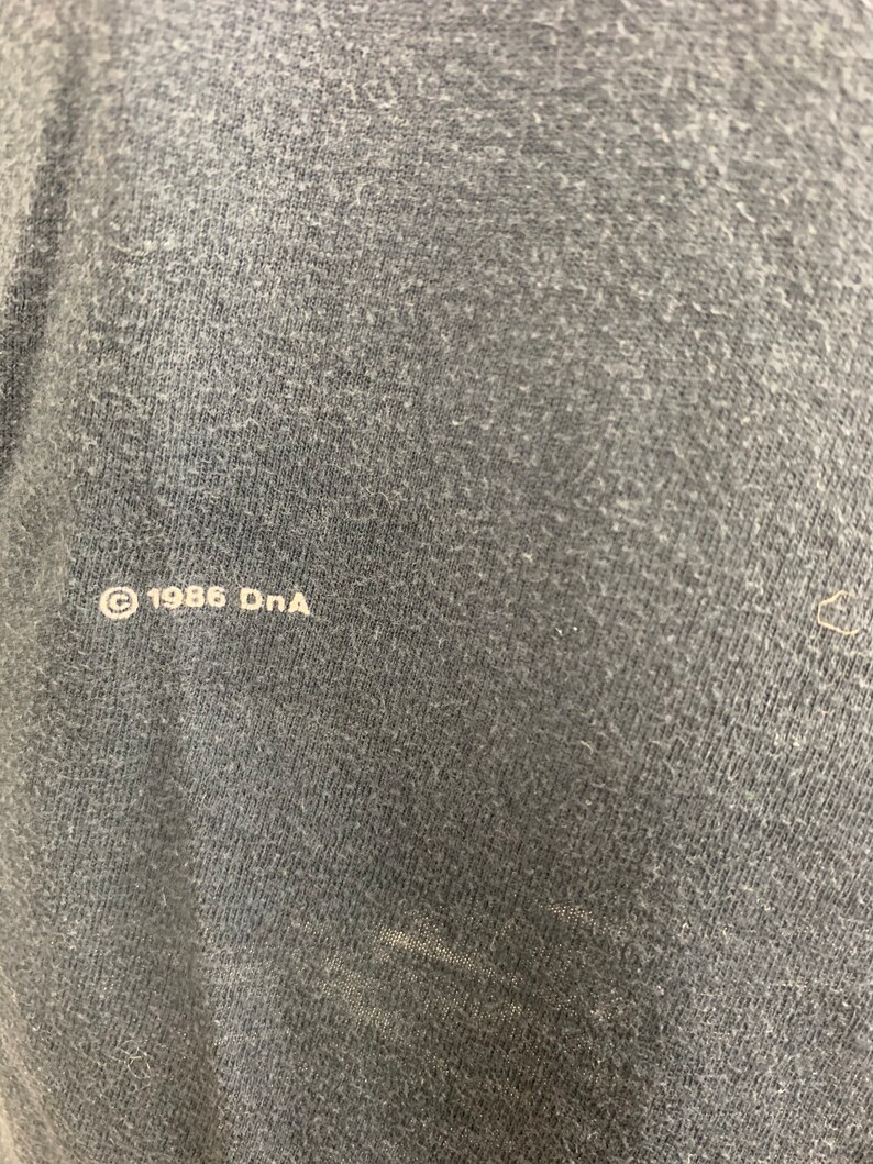 May include: Close-up of a dark grey fabric with the text "&copy; 1986 DnA" printed in white.