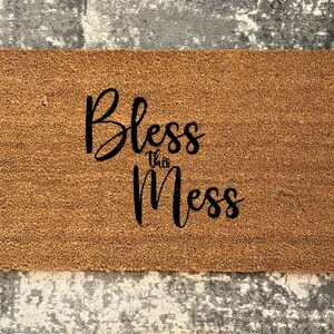 Puede incluir: Una alfombra rectangular hecha de fibras naturales de coco. La alfombra presenta la frase "Bless this Mess" en una fuente cursiva negra. La alfombra está colocada sobre una alfombra gris y blanca estampada.
