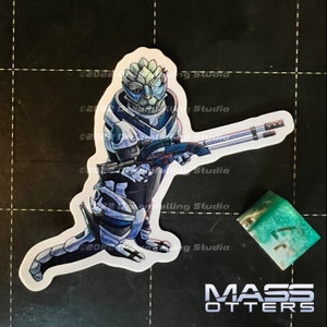 May include: A sticker featuring a blue and white space suit character holding a gun. The character has green skin and a blue helmet. The sticker is on a black background with the text "MASS OTTERS" below it.