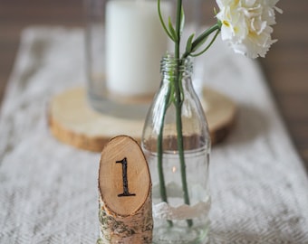 May include: A wooden table number with the number "1" on it, sitting on a table runner with a glass vase filled with pink and white carnations.