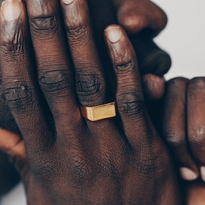 Op de afbeelding: Een close-up van een hand met een gouden vierkante ring. De hand rust op het gezicht en de vingers zijn zichtbaar.