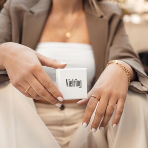 May include: A white box with the word "Vielring" printed on it. The box is being held by a person wearing a brown jacket and a white top.