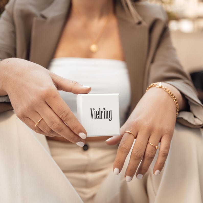 May include: A white box with the word "Vielring" printed on it. The box is being held by a person wearing a brown jacket and a white top.