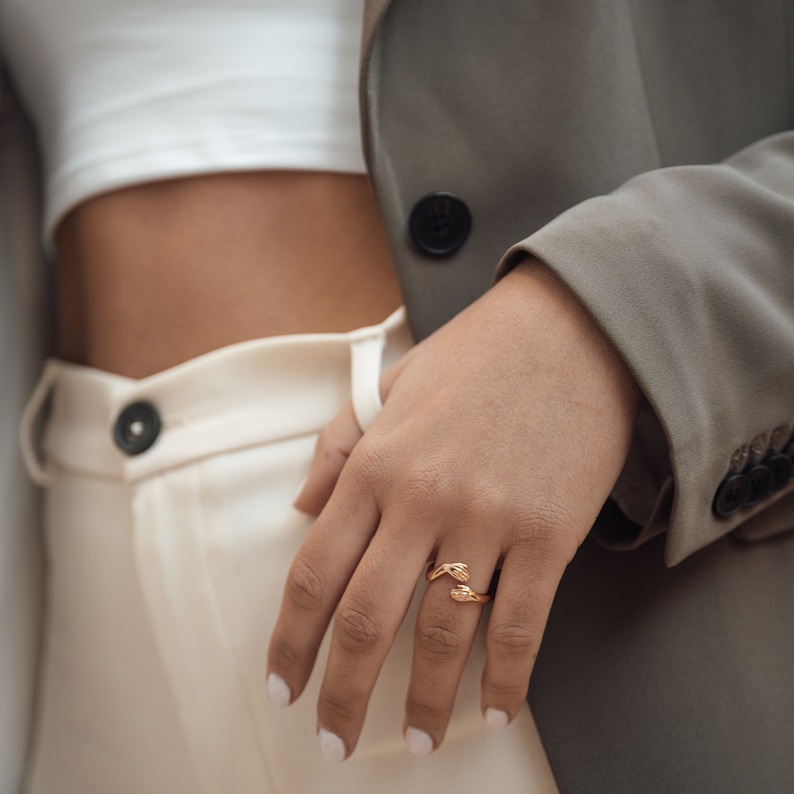 Pu&ograve; includere: Un anello d'oro con un design di due mani intrecciate. L'anello &egrave; indossato sull'anulare di una mano.