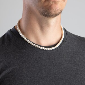 May include: A white pearl necklace on a person's neck. The necklace is made of small, round pearls and is worn on a dark grey t-shirt.