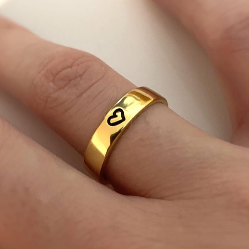 May include: A gold band ring with a small heart design engraved on the top.