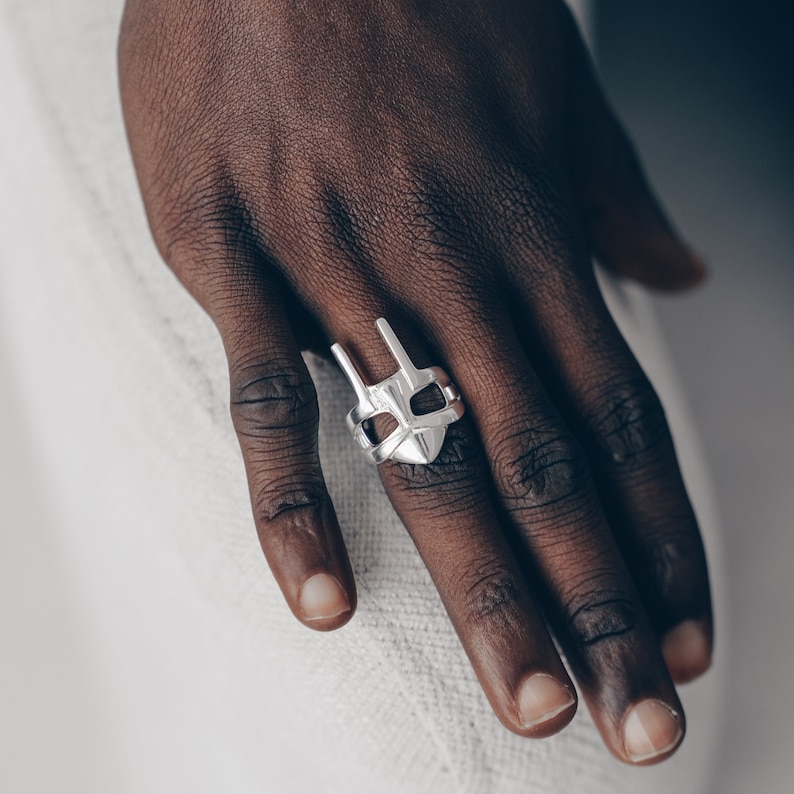 Peut inclure: Une bague en argent avec un motif g&eacute;om&eacute;trique ressemblant &agrave; un masque. La bague est port&eacute;e &agrave; l'index d'une main.