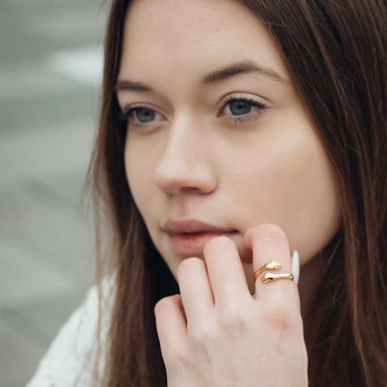 以下が含まれることがあります： 2つの手が抱き合うデザインのゴールドカラーのリング。指に装着され、手が指を包み込んでいます。リングは滑らかで光沢のある仕上がりです。