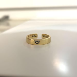 May include: A gold adjustable ring with the words "LOVE" and "YOU" engraved on the outside and a heart symbol engraved on the inside.