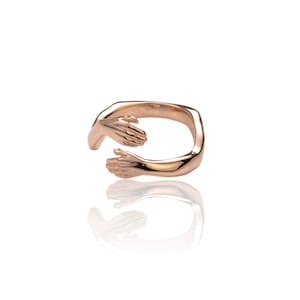 Pu&ograve; includere: Un anello in oro rosa con due mani intrecciate in un abbraccio. L'anello &egrave; in metallo e ha una finitura liscia.