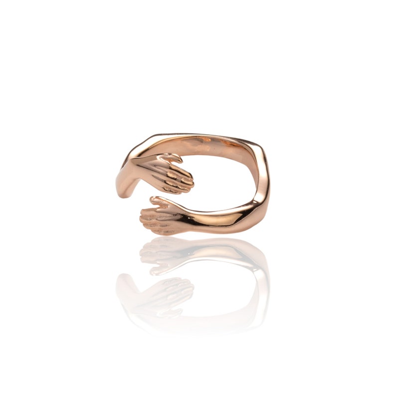 Pu&ograve; includere: Un anello in oro rosa con due mani intrecciate in un abbraccio. L'anello &egrave; in metallo e ha una finitura liscia.