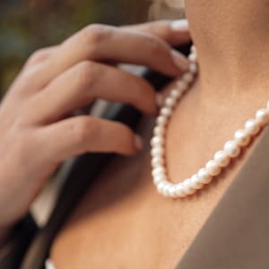 May include: A woman wearing a pearl necklace. The pearls are white and round. The necklace is simple and elegant.