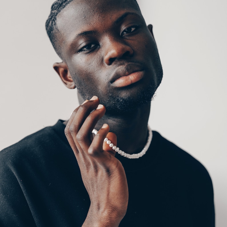 May include: A close-up portrait of a person with dark skin wearing a black shirt and a white pearl necklace. The person is looking directly at the camera with a serious expression.