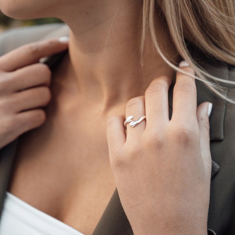 Pu&ograve; includere: Un anello d'argento con un designo unico che presenta due mani intrecciate. L'anello &egrave; indossato sul dito di una mano.