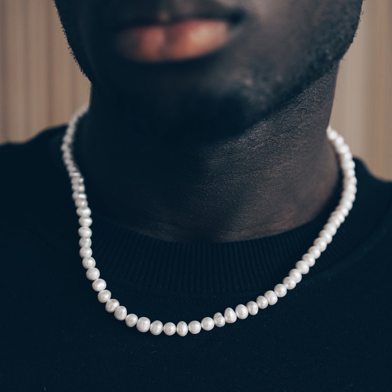 May include: A white pearl necklace on a person's neck. The necklace is made of small, round pearls that are strung together on a thin chain.
