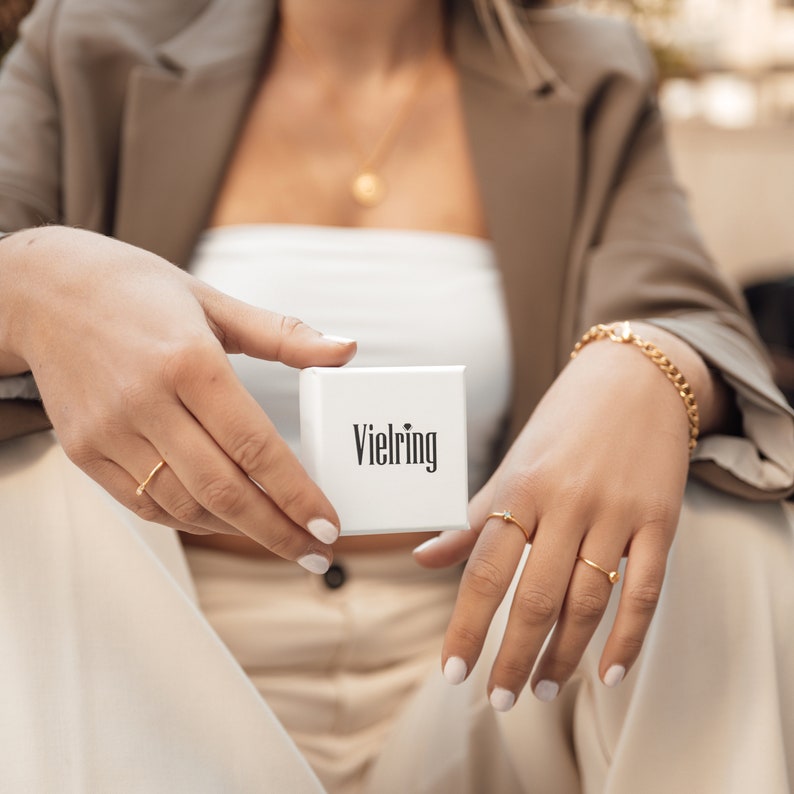 May include: A white box with the word "Vielring" printed on it. The box is being held by a person wearing a brown jacket and a white top.