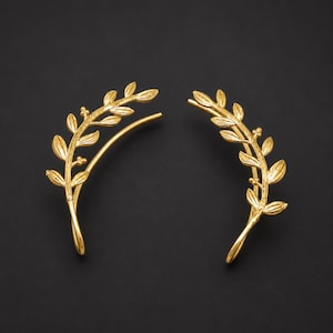 May include: A pair of gold-toned ear climbers shaped like curved olive branches. Each piece features a series of small, textured leaves and tiny spherical berries arranged along a slender, arching stem that follows the contour of the earlobe.