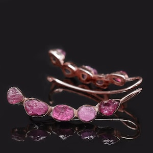 Op de afbeelding: Een paar roségouden oorcrawlers, elk met vijf ruwe, roze edelstenen. De oorbellen volgen de vorm van het oor, met een pin- en haakontwerp. De achtergrond is een donker, reflecterend oppervlak.