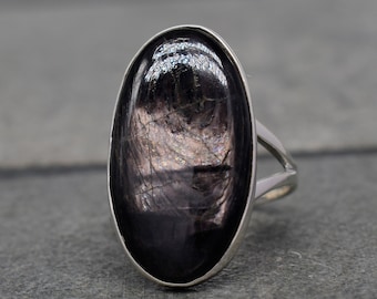Puede incluir: Un anillo de plata con una gran piedra preciosa negra de forma ovalada. La piedra preciosa tiene un efecto brillante e iridiscente.