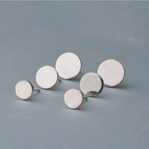 May include: Five silver circle stud earrings of varying sizes on a light blue background.
