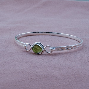 Peut inclure: Un bracelet jonc en argent avec une texture martelée. Le bracelet présente une pierre précieuse brute vert clair au centre, maintenue par une monture décorative en argent. Le bracelet est présenté sur une surface rose.