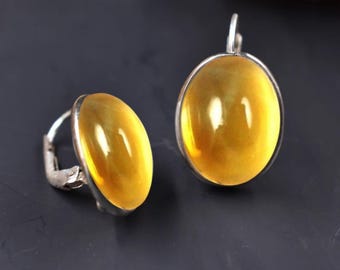 May include: A pair of earrings featuring smooth, oval-shaped yellow amber stones set in polished silver metal. Each earring has a secure lever-back closure mechanism, showcasing the translucent, warm golden hue of the polished amber cabochons.