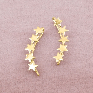 Puede incluir: Un par de pendientes de estrella de oro. Cada pendiente tiene una fila de siete estrellas doradas dispuestas en una línea curva.