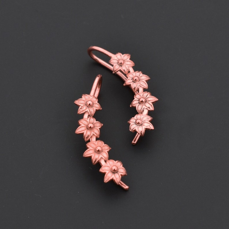 Pode incluir: Um par de brincos trepadores florais em ouro rosa. Cada trepador apresenta uma s&eacute;rie de flores detalhadas. Os brincos s&atilde;o projetados para curvar-se na orelha, oferecendo um estilo &uacute;nico e elegante. O fundo &eacute; cinza escuro s&oacute;lido.