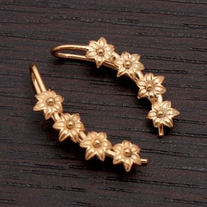 Pode incluir: Dois brincos trepadores dourados, cada um com uma s&eacute;rie de pequenos desenhos florais detalhados. Os brincos curvam-se para se ajustarem &agrave; orelha, com um pino numa extremidade e um gancho na outra. Apresentados numa superf&iacute;cie de madeira escura.