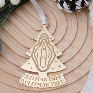 May include: A gold-colored Christmas tree ornament with the words "Clitmas Tree O Clitmas Tree". The ornament is shaped like a Christmas tree with a stylized vulva design. The ornament hangs from a white ribbon.