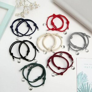 Puede incluir: Surtido de pulseras de cuerda a juego en varios colores, incluyendo rojo, negro y crema. Cada pulsera presenta un diseño de nudo y un pequeño dije redondo. Las pulseras se exhiben sobre una superficie blanca.