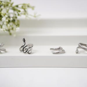 May include: Four silver snake rings on a white surface. The rings feature detailed snake scale textures and various coiled designs. White flowers are in the background.