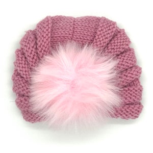 May include: A mauve knitted baby turban featuring a large, fluffy pink pom-pom. The turban has a ribbed design and is made of soft, textured yarn. This stylish headwear is suitable for infants and young children.