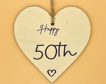 Puede incluir: Adorno de madera en forma de corazón color crema con las palabras "Happy 50th" escritas en negro. Un pequeño corazón está dibujado debajo del número 50. Tiene una cuerda de yute para colgar, ideal para un 50 cumpleaños.