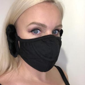 Könnte beinhalten: Schwarze Gesichtsmaske mit angebrachten Kunstfell-Ohrenschützern. Die Maske besteht aus einem weichen, dunklen Stoff und hat ein konturiertes Design. Die Ohrenschützer sind aus einem dunklen, flauschigen Material. Die Person hat blonde Haare und blaue Augen.