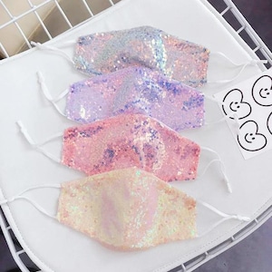 May include: Four sequined face masks in pastel colors: iridescent, lavender, pink, and peach. Each mask has white adjustable ear loops. The masks are arranged on a white surface, with a small sticker sheet with cartoon faces.