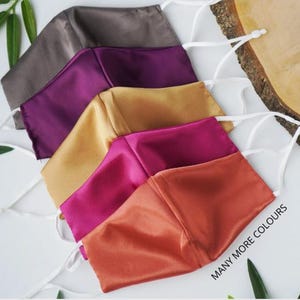 Peut inclure: Cinq masques faciaux de couleurs variées : marron, violet, doré, fuchsia et orange. Chaque masque est doté d'élastiques blancs réglables. Le texte "MANY MORE COLOURS" est visible en bas à droite.