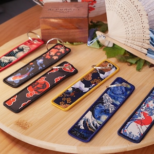 K&ouml;nnte beinhalten: Eine Sammlung von Schl&uuml;sselanh&auml;ngern im japanischen Stil mit verschiedenen Designs, darunter Wellen, Koi-Fische und Kraniche. Jeder Schl&uuml;sselanh&auml;nger ist rechteckig mit einem Metallring und einem farbenfrohen Stoffdesign. Sie sind auf einem Holztablett angeordnet.