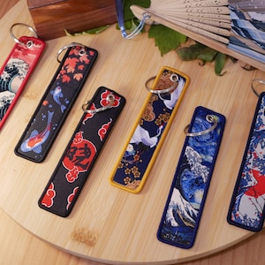 K&ouml;nnte beinhalten: Bestickte Schl&uuml;sselanh&auml;nger mit japanisch inspirierten Designs. Die Schl&uuml;sselanh&auml;nger zeigen verschiedene Muster, darunter Wellen, Koi-Fische, Kraniche und florale Motive. Jeder Schl&uuml;sselanh&auml;nger hat einen Metallring zur Befestigung und ist rechteckig.