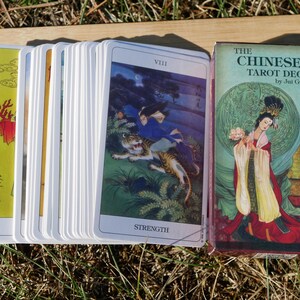 May include: A deck of tarot cards with a box. The cards feature colorful illustrations of Chinese art. The box reads "THE CHINESE TAROT DECK" by Jui Guoliang. The cards are fanned out, showing the "Strength" card with a tiger.
