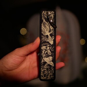 K&ouml;nnte beinhalten: Schwarzes rechteckiges Etui mit einem goldenen Koi-Fisch-Design. Das Etui hat einen Metallring zur Befestigung. Die Koi-Fische schwimmen in einem detaillierten, flie&szlig;enden Muster. Der Hintergrund ist dunkel, mit verschwommenen Lichtern.